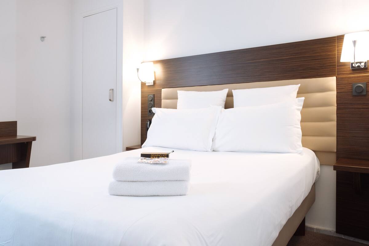 A neatly made bed with white linens, a small bedside table with a book, and a wall-mounted lamp.