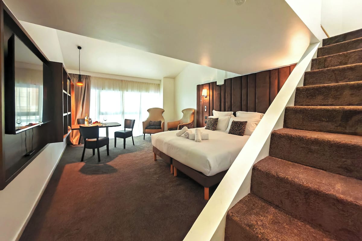 A modern hotel room with a large bed, a dining area, and a staircase.