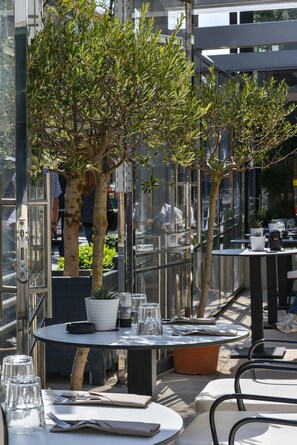 Un área de comedor al aire libre con mesas y sillas, plantas en macetas y una barandilla de cristal.