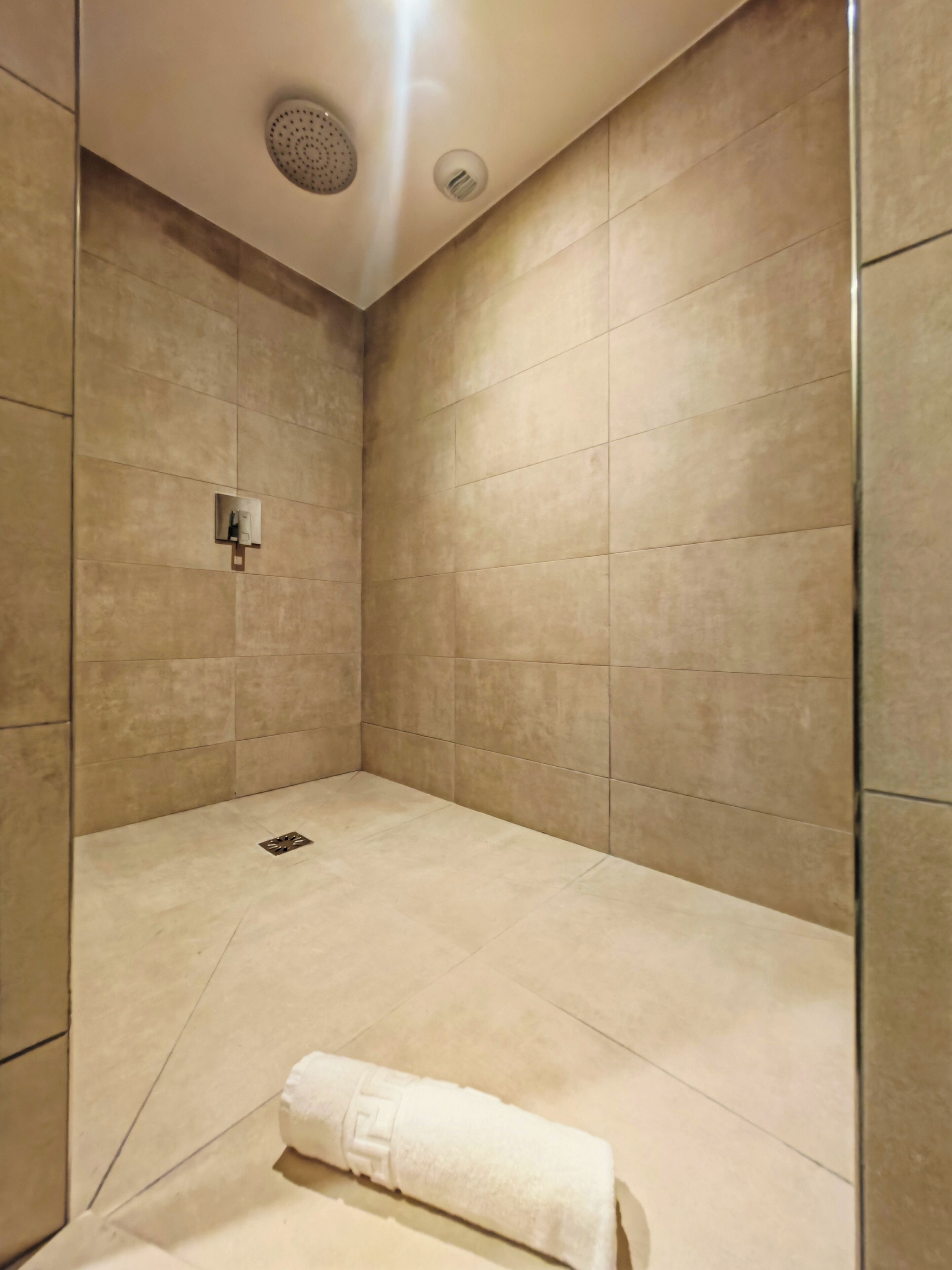 A modern bathroom with beige tiles, a showerhead, a towel, and a light switch.