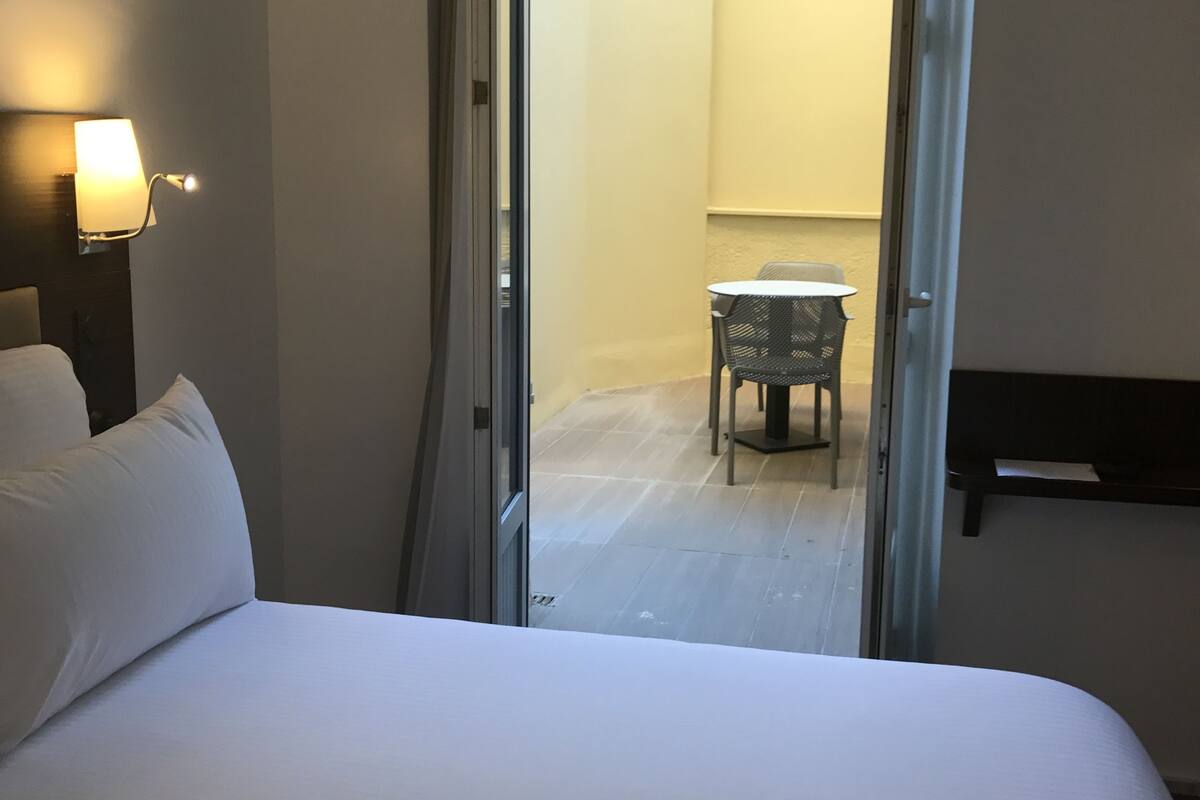 A hotel room with a bed, a nightstand with a lamp, and a view of an outdoor area with a table and chairs.