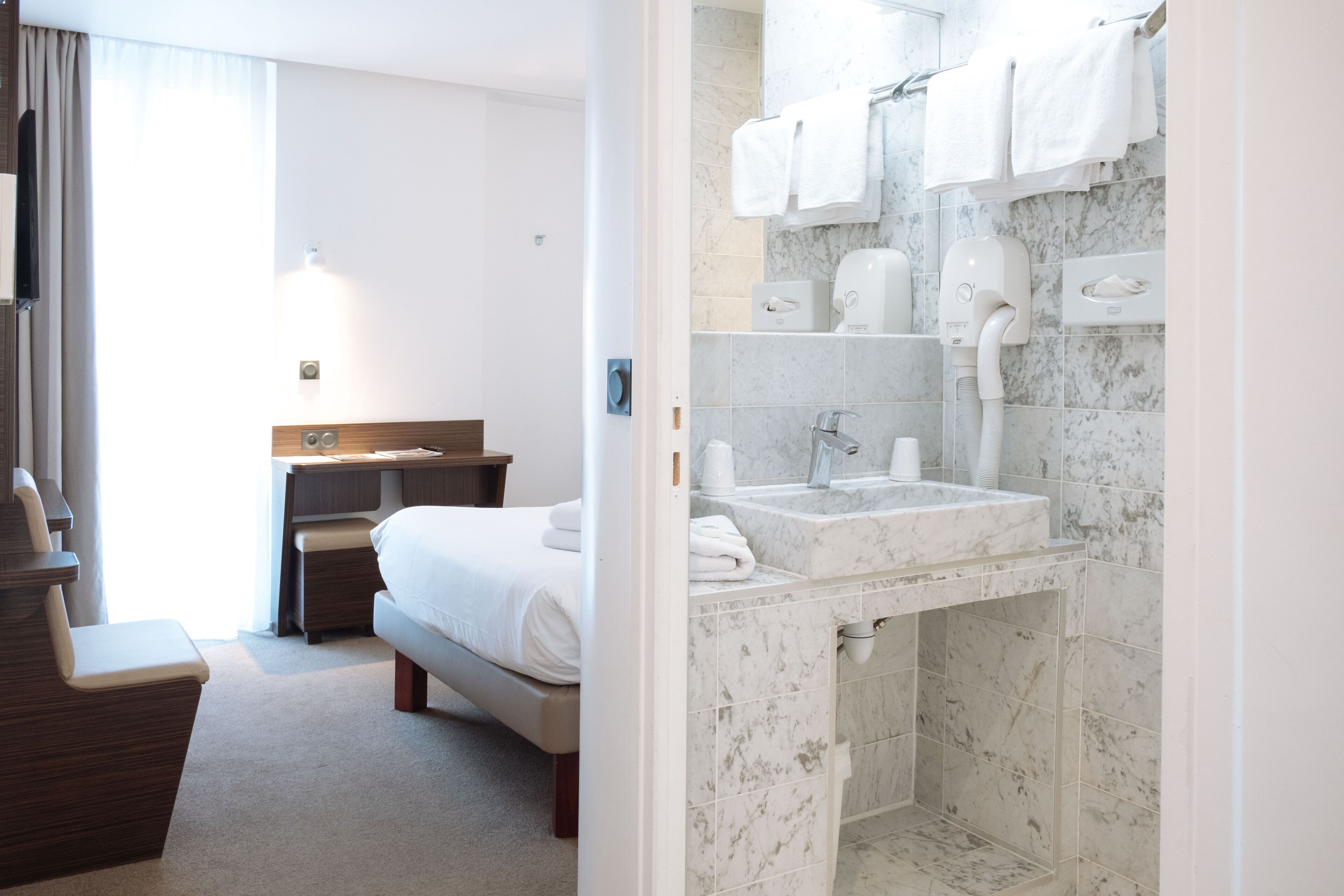A hotel room with a bed, a desk, a bathroom with a sink and towel dispenser.