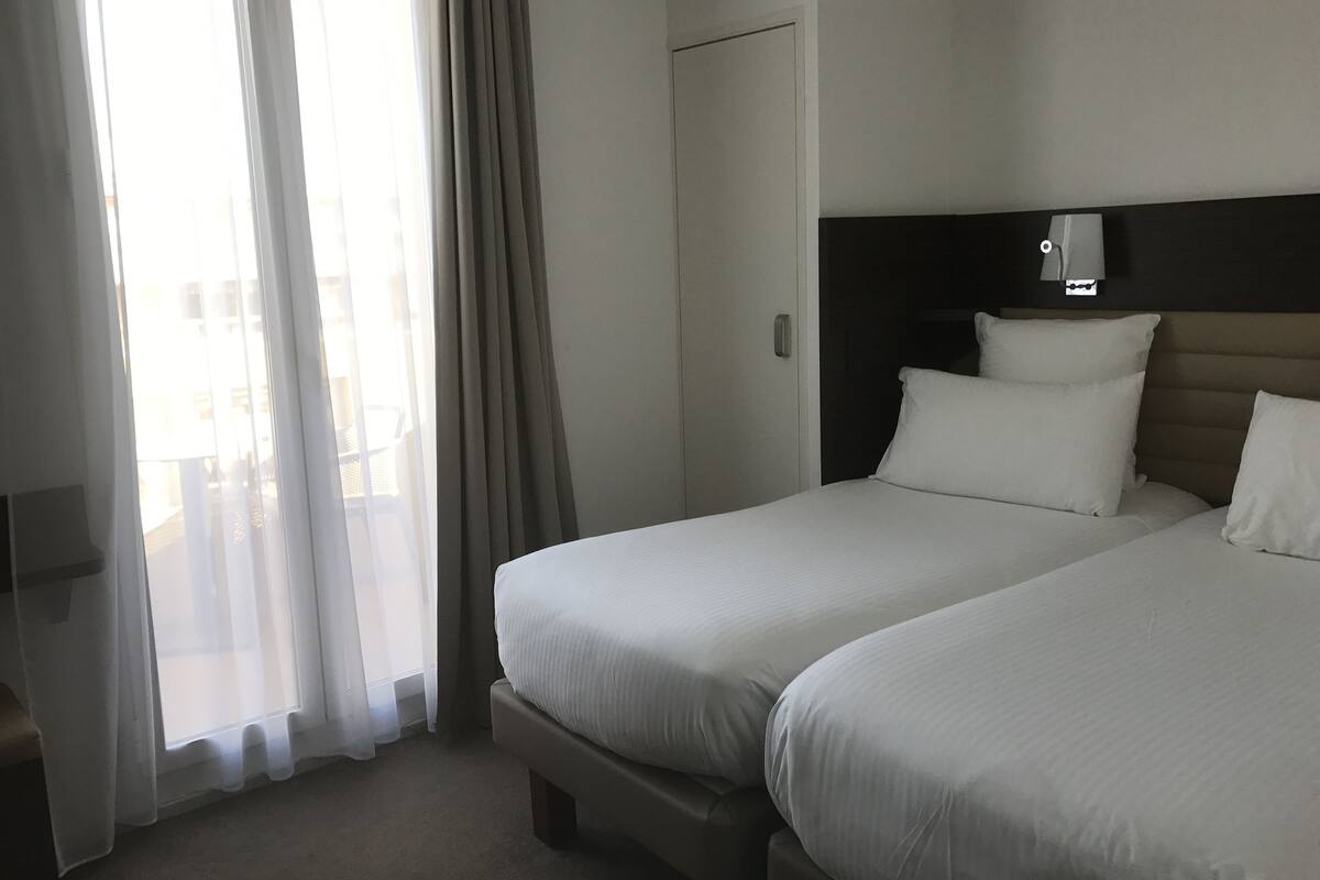 A hotel room with two beds, a headboard with a light switch, a window with sheer curtains, and a view of buildings outside.