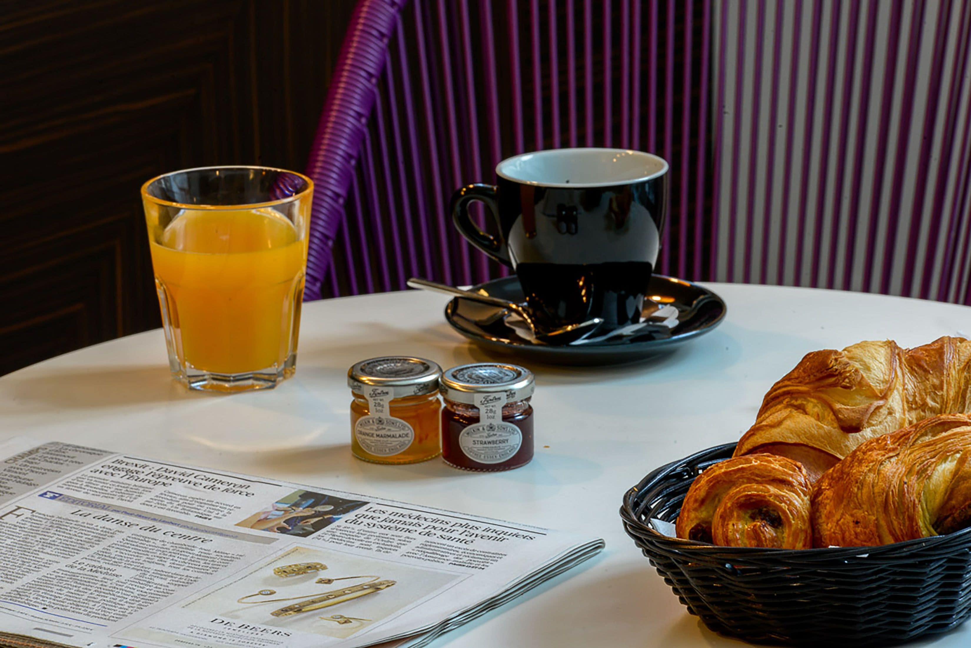 Une table de petit-déjeuner avec des croissants, un verre de jus d’orange, une tasse de café et deux pots de confiture.