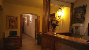 Un espace d’accueil d’hôtel avec un comptoir en bois, un ordinateur et une chaise. Un couloir mène à une pièce où se trouvent un tableau et une plante en pot.