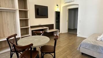 Een compacte kamer met een ronde tafel en vier stoelen, een bed, een aan de muur bevestigde televisie, en een garderobe met open planken.