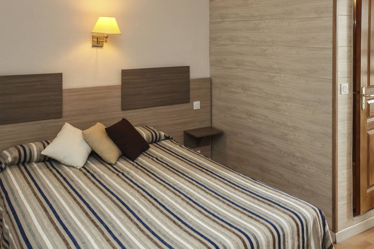 A hotel room with a bed, a nightstand, a wall-mounted lamp, and a wooden door.