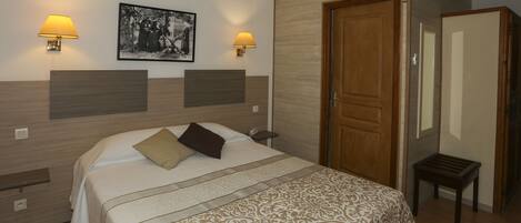 A hotel room with a bed, two wall-mounted lamps, a framed picture, a wooden door, and a small table.