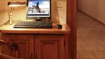 A wooden desk with a computer, a lamp, and a chair in a hallway with tiled flooring and a framed picture on the wall.