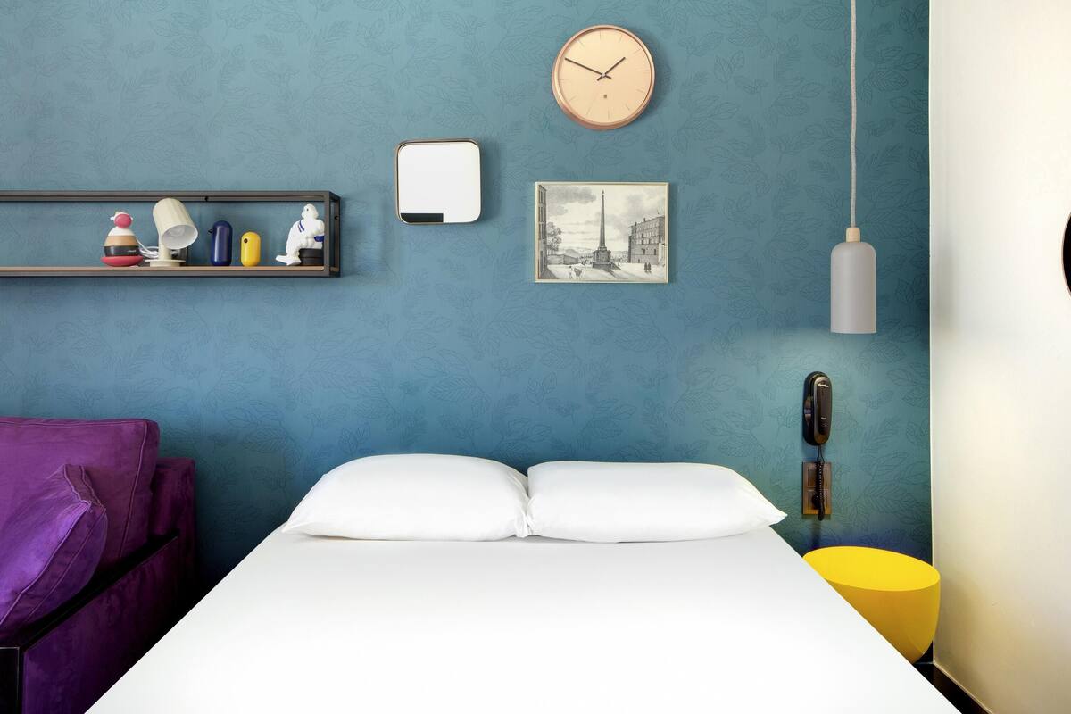 Una habitación con una cama, un reloj, un estante con adornos, un cuadro y una lámpara colgante.