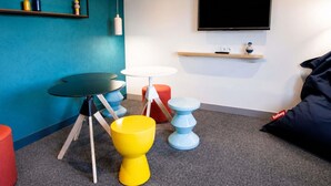 Una habitación con pared verde azulada, un televisor de pantalla plana, una mesa redonda blanca, un taburete amarillo y un taburete azul.