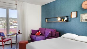 A hotel room with a bed, a purple sofa, a small table with a toy, a chair, and a view of the city through a large window. - Hôtel ibis Styles Clermont-Ferrand Gare (Clermont-Ferrand)