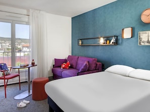 A hotel room with a bed, a purple sofa, a small table with a toy, a chair, and a view of the city through a large window. - Hôtel ibis Styles Clermont-Ferrand Gare (Clermont-Ferrand)