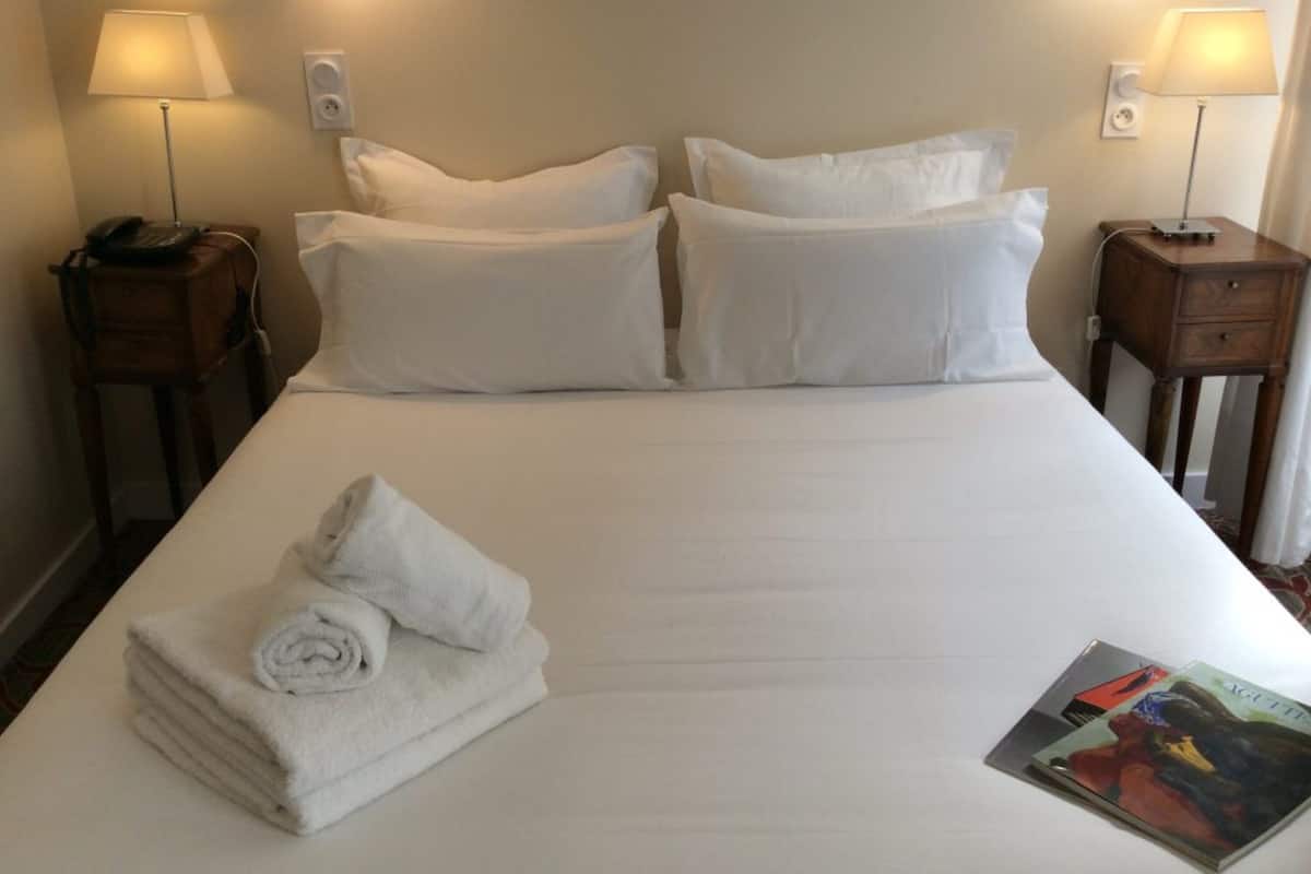 Una cama bien tendida con sábanas blancas, dos mesitas de noche con lámparas y una toalla doblada sobre la cama.