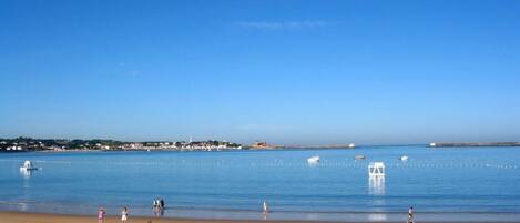 A beach with people sunbathing and swimming, boats on the water, and a clear sky.