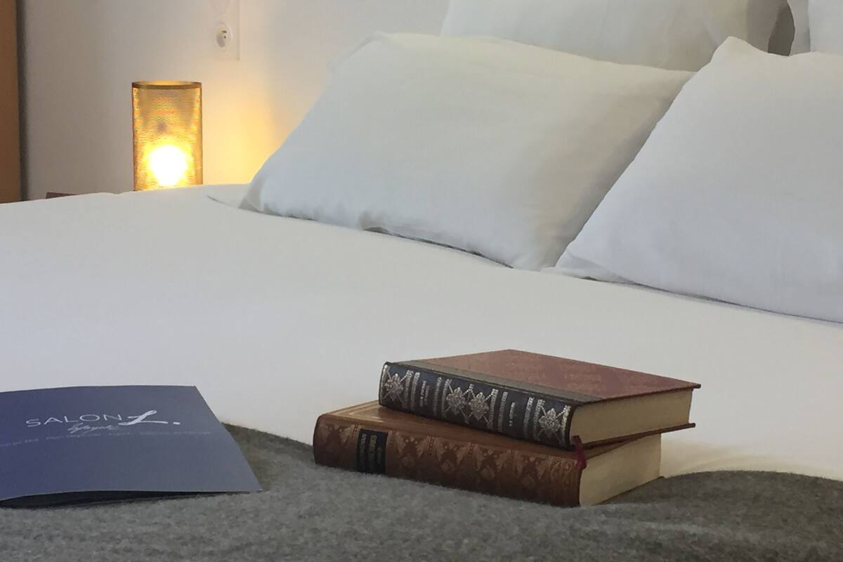 Una cama bien hecha con sábanas y almohadas blancas, una lámpara en la mesita de noche y dos libros sobre la cama.