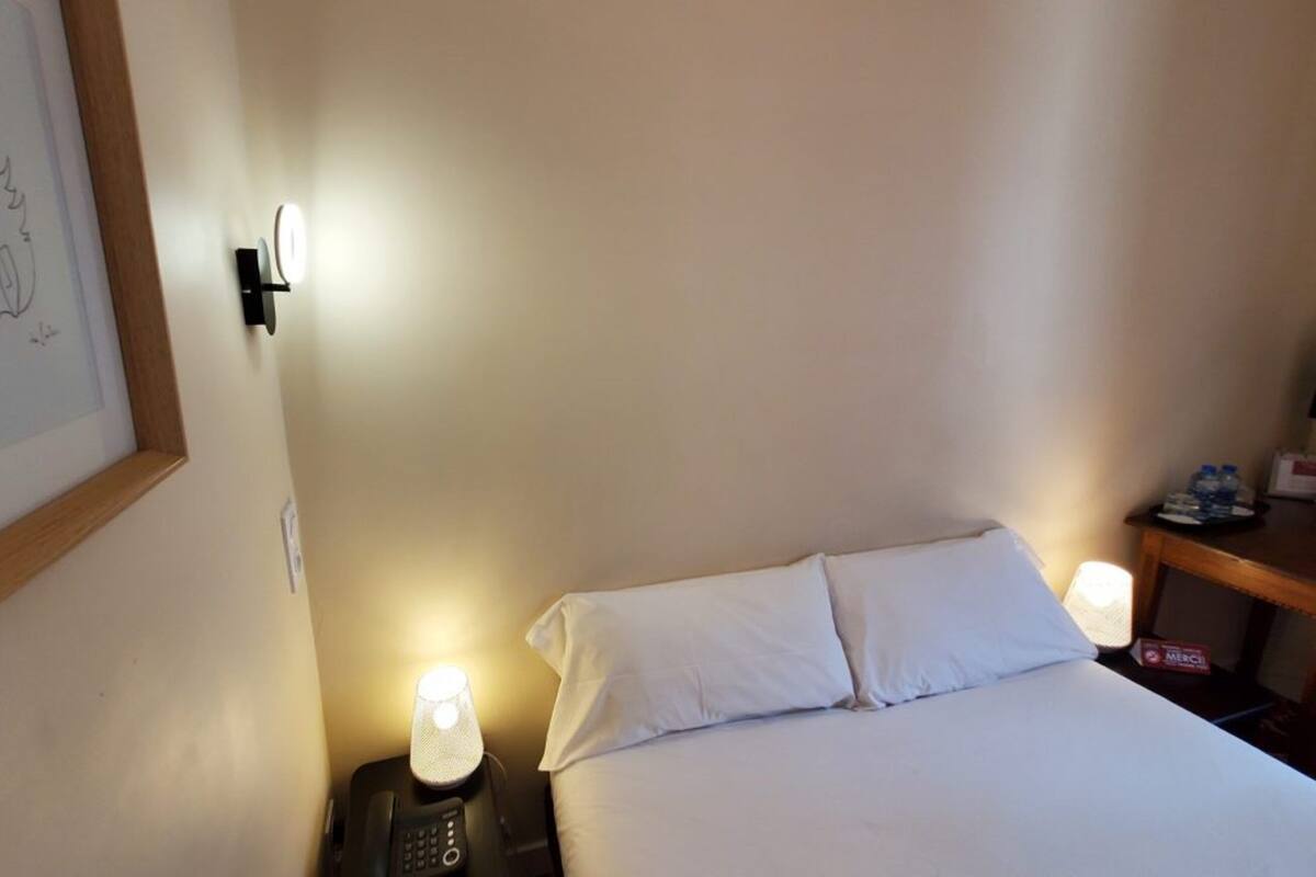 Habitación de hotel con cama, lámparas de noche, mesita de noche, un dibujo enmarcado en la pared y una pequeña mesa con botellas de agua.