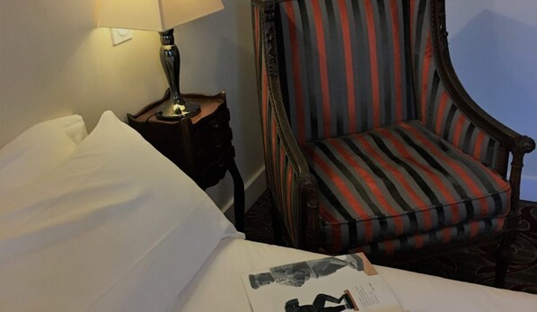 A hotel room with a neatly made bed, a striped armchair, a small table with a lamp, and a wall socket.