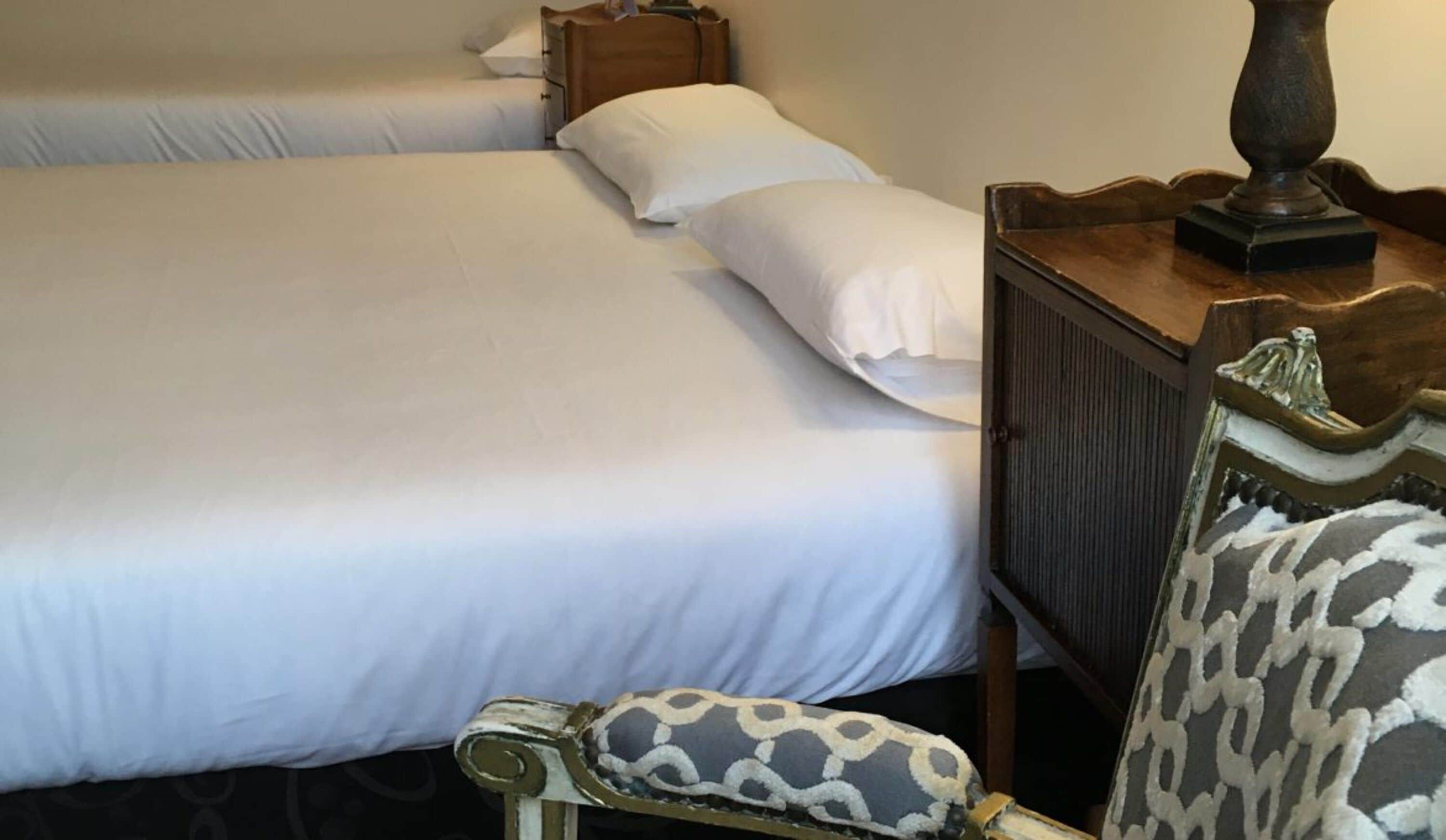 A hotel room with two beds, a wooden nightstand, and a patterned armchair.