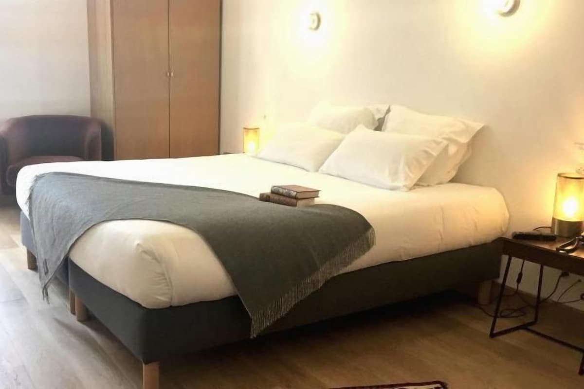 Un lit avec une tête de lit sombre, une table de chevet avec une lampe et une chaise dans une pièce dont le sol est en bois.