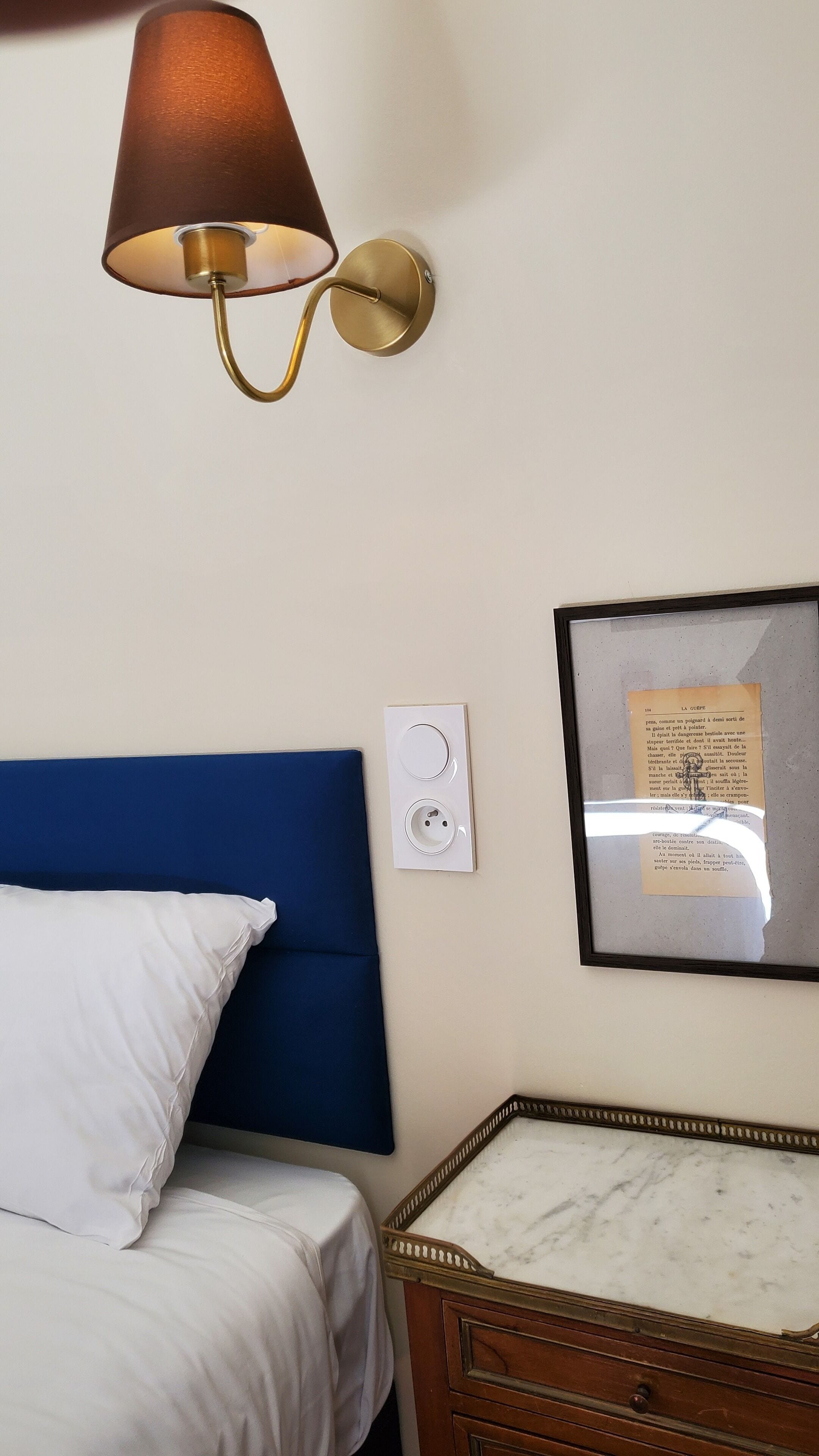 Ein Schlafzimmer mit einem Bett, einer Wandlampe, einer Steckdose und einem gerahmten Bild an der Wand.