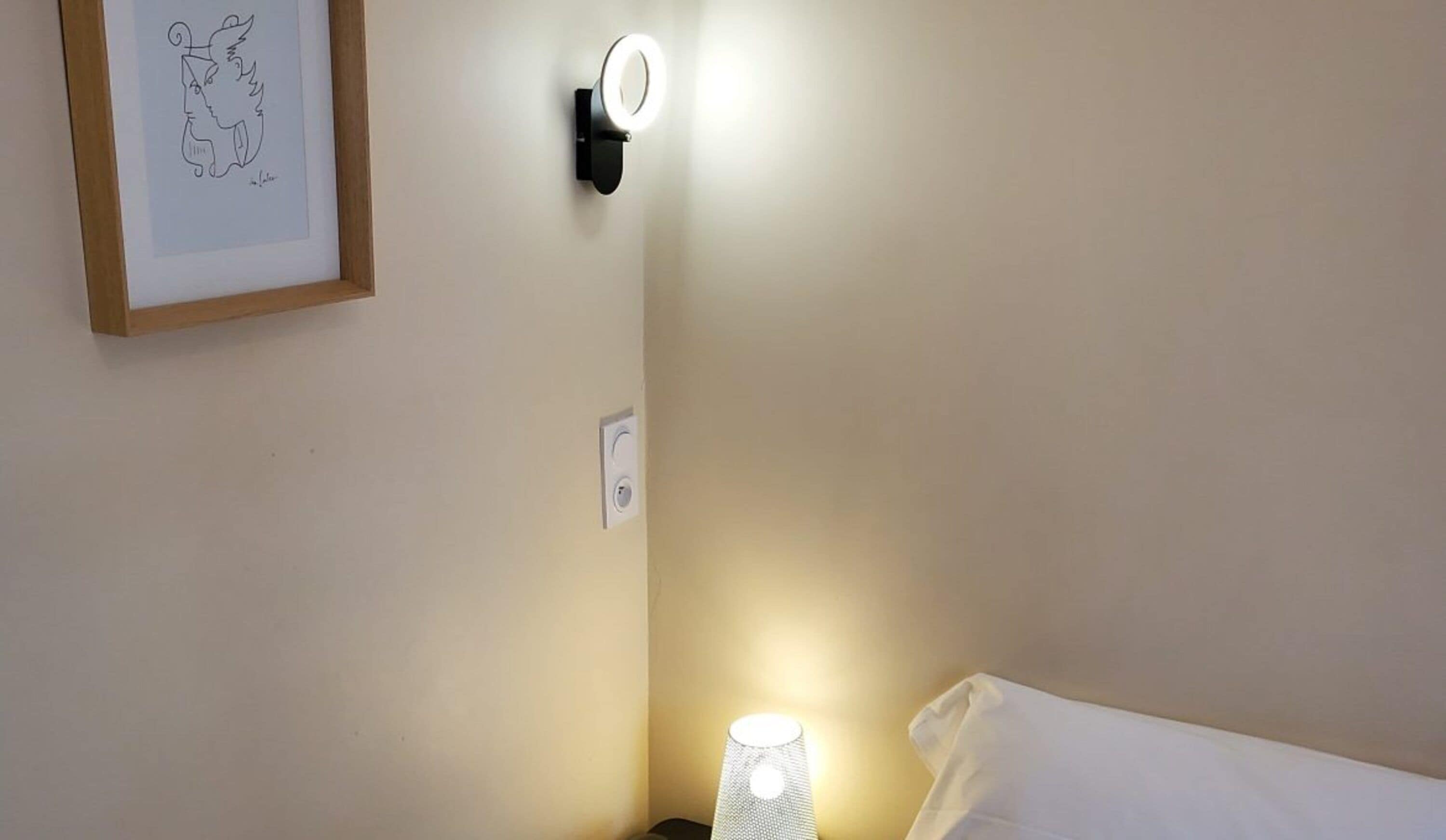 Una habitación de hotel con una lámpara de noche, una obra de arte enmarcada en la pared y un aplique.