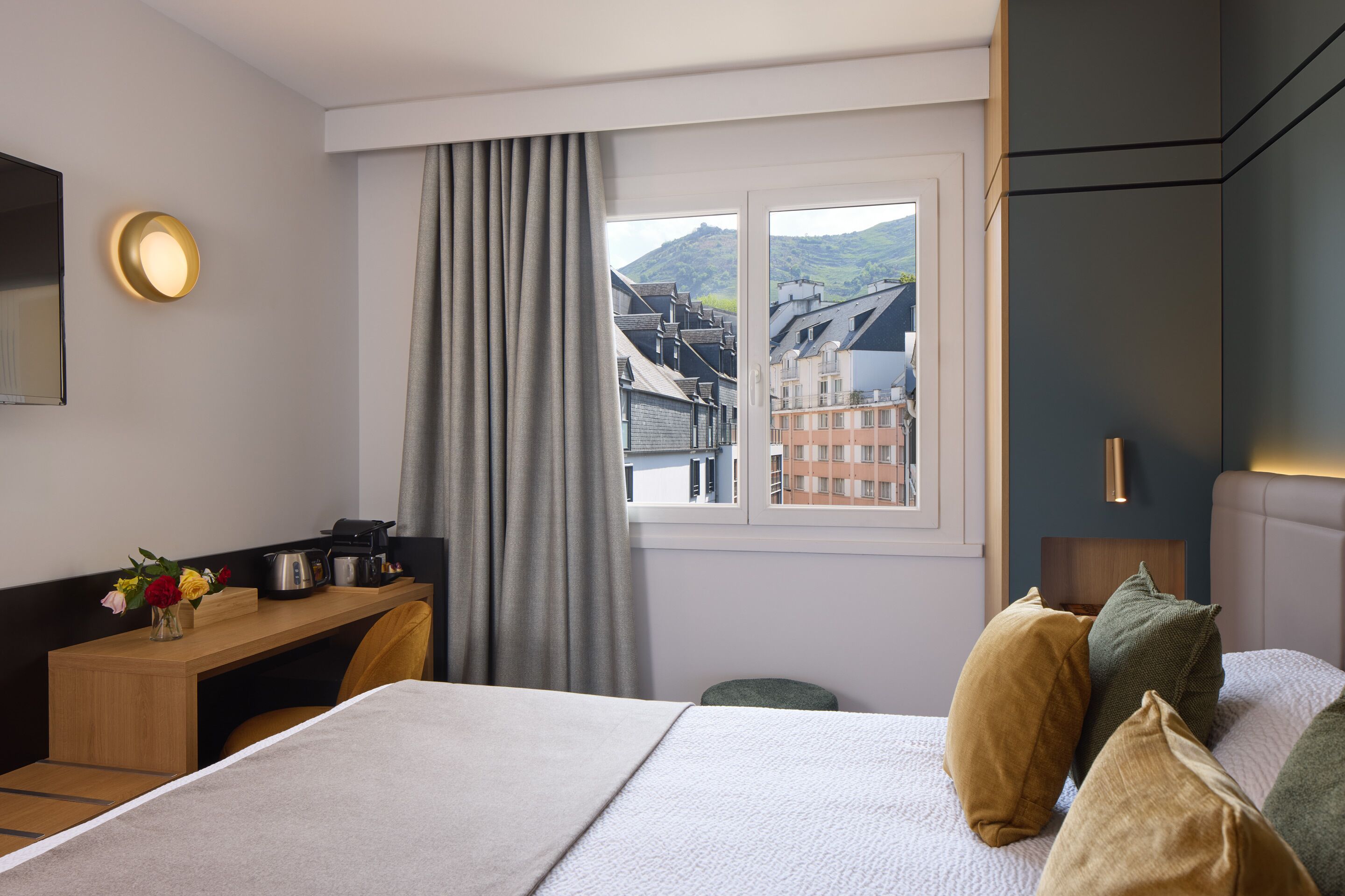 Une chambre d’hôtel avec un lit, un bureau doté d’une machine à café, une vue sur les bâtiments par la fenêtre et une table de chevet avec une lampe.