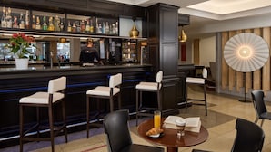 A bar area with stools, a bar counter with liquor bottles, and a table with a croissant and a book. - Hotel Roissy Lourdes (Lourdes)