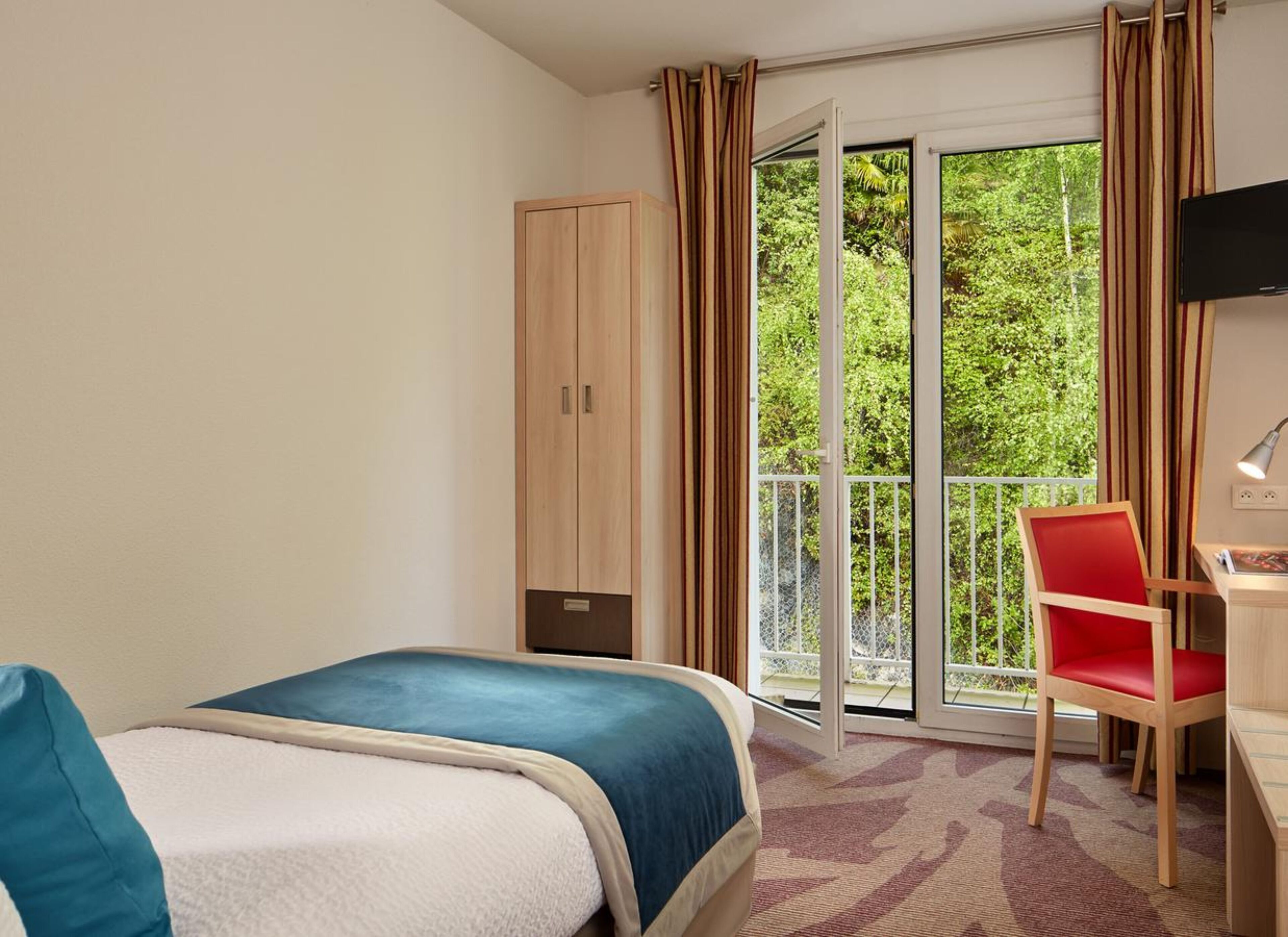 A hotel room with a bed, a desk with a chair, a wardrobe, and a balcony with curtains.