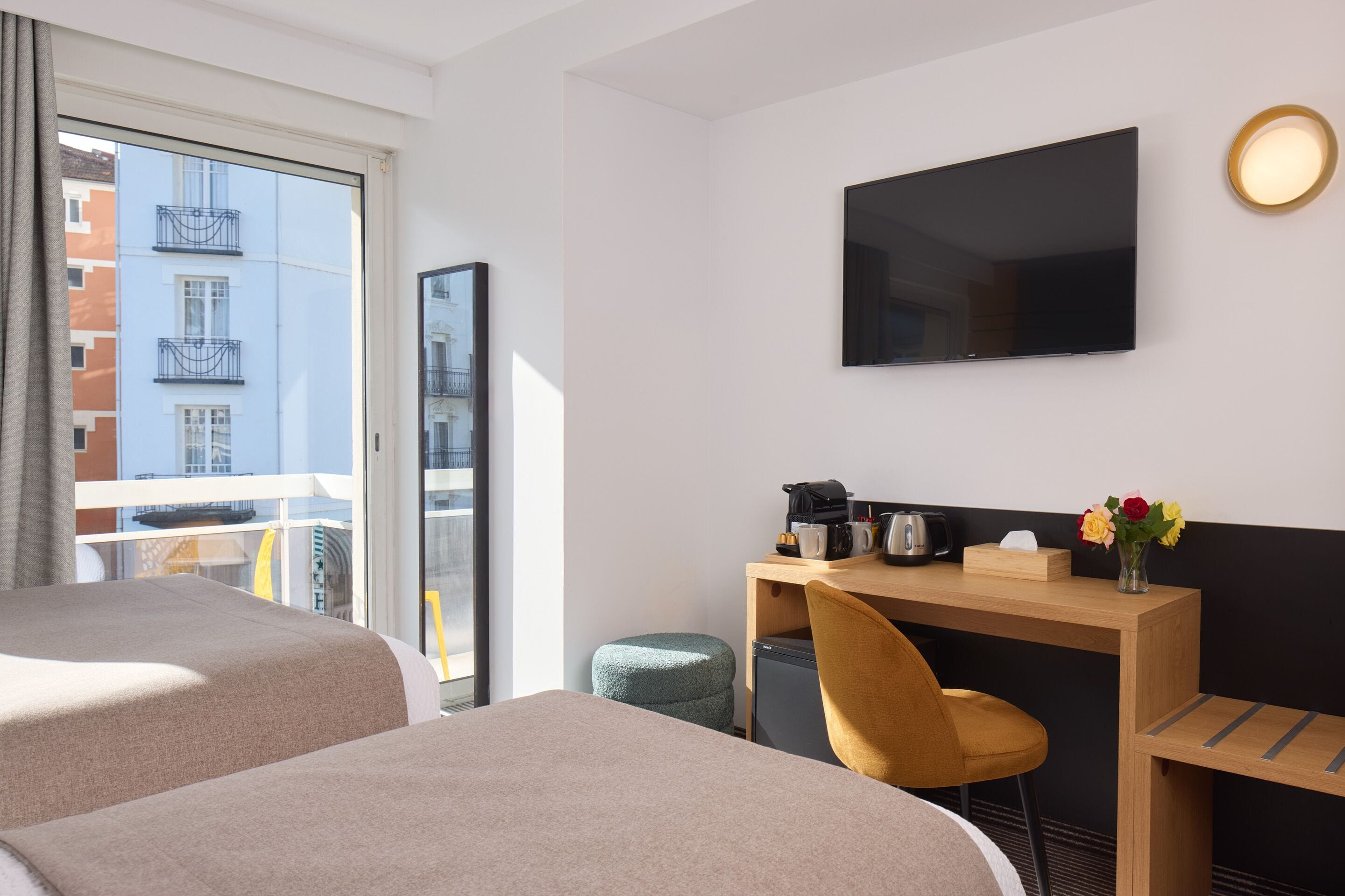 A hotel room with two beds, a desk with a chair, a television, and a window with a view of buildings.