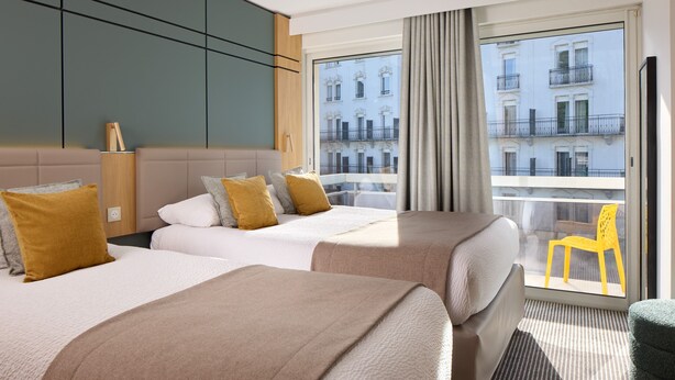 A hotel room with two beds, a large window with a view of buildings, a chair, and a small table. - Hotel Roissy Lourdes (Lourdes)