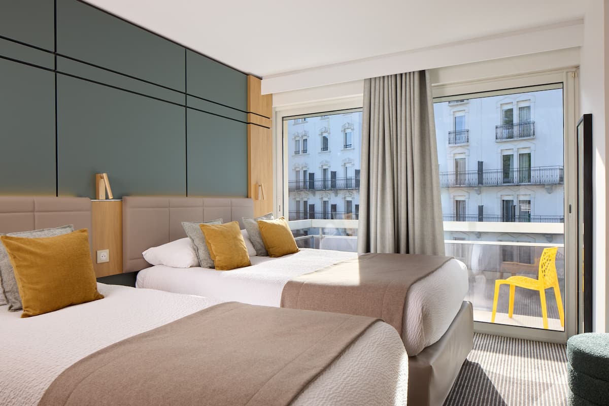 A hotel room with two beds, a large window with a view of buildings, a chair, and a small table.