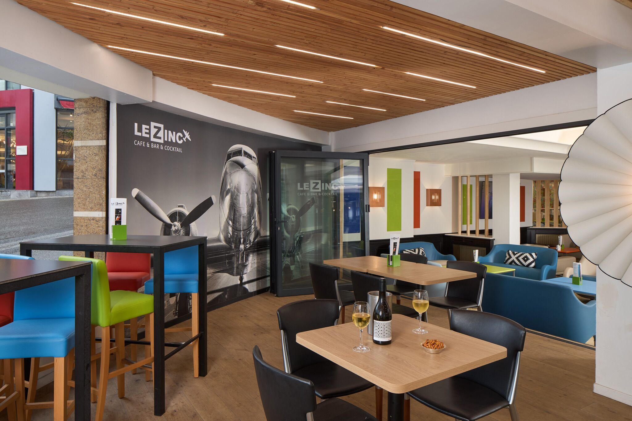Ein modernes Café mit Holzbodenpaneelen, bunten Stühlen und einem gut sichtbaren LEZINC-Logo an der Wand.