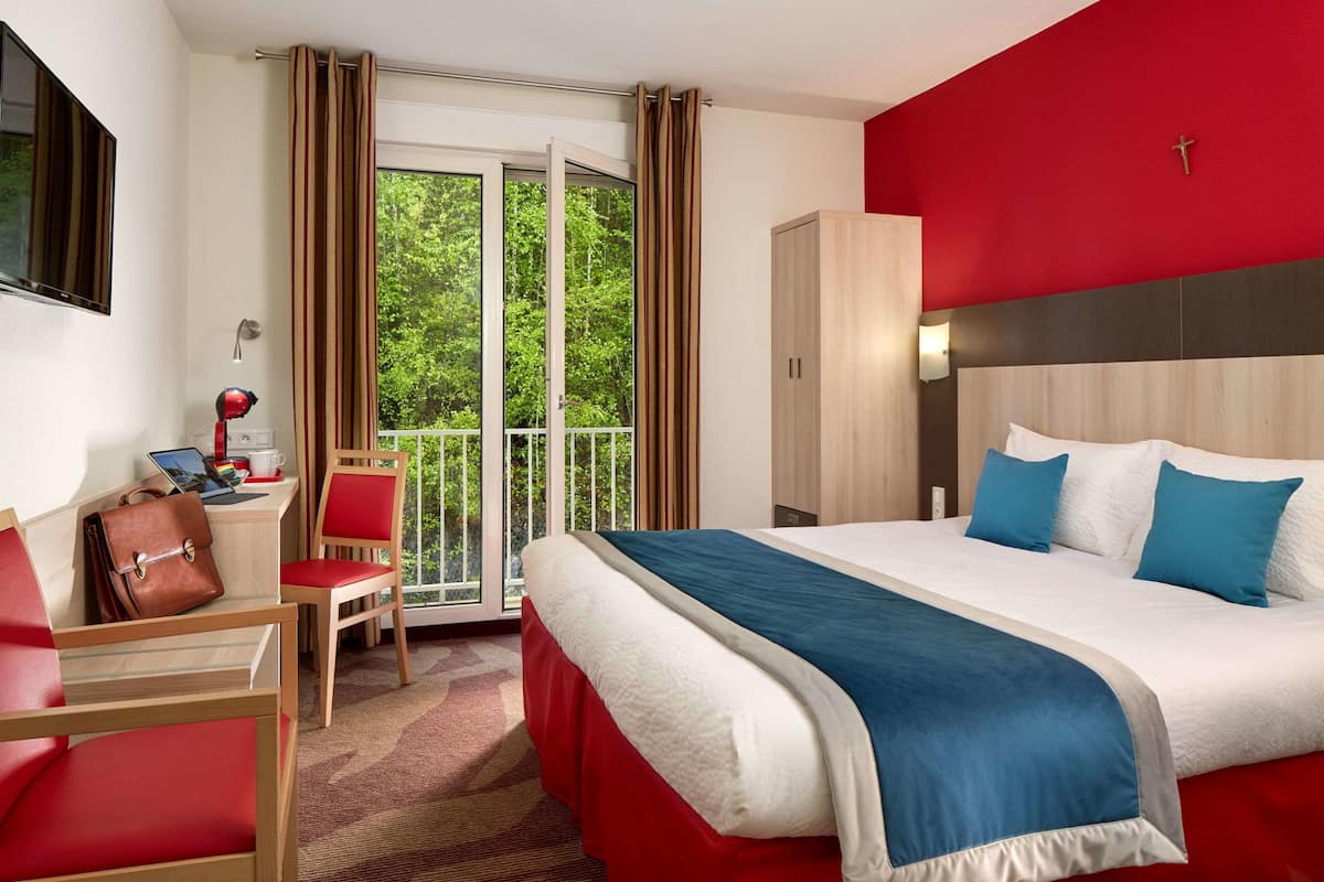 A hotel room with a bed, a desk with a laptop, a chair, and a view of greenery through the window.