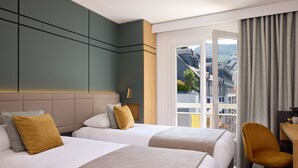 A hotel room with two beds, a desk, a chair, and a view of the city. - Hotel Roissy Lourdes (Lourdes)
