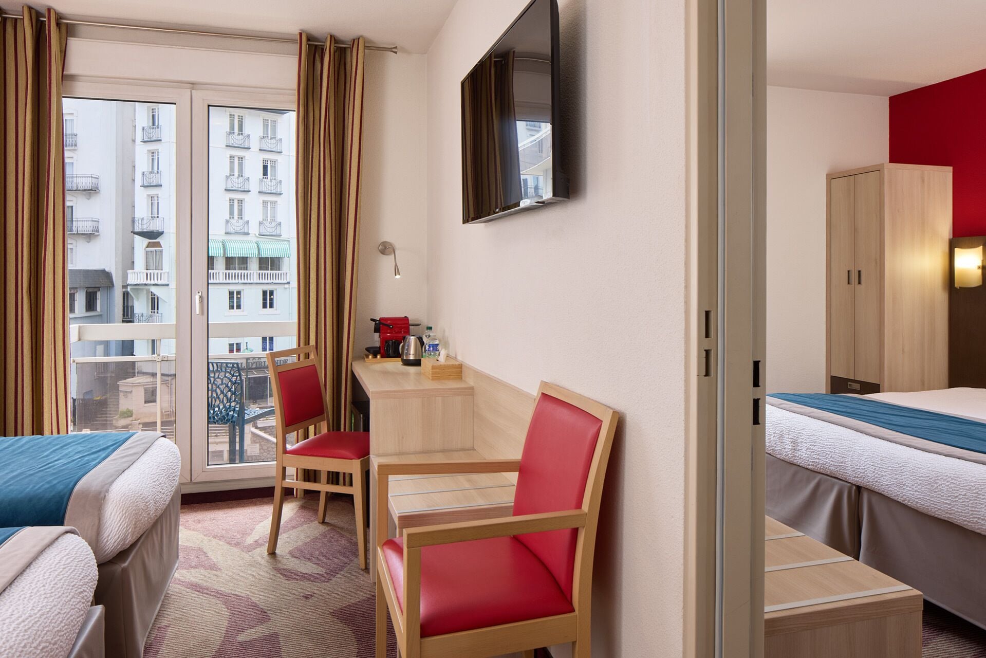 Een hotelkamer met twee bedden, een bureau met stoel, een aan de muur bevestigde televisie en uitzicht op de stad door het raam.