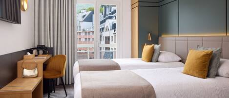 A hotel room with two beds, a desk, a chair, and a view of buildings through a window.
