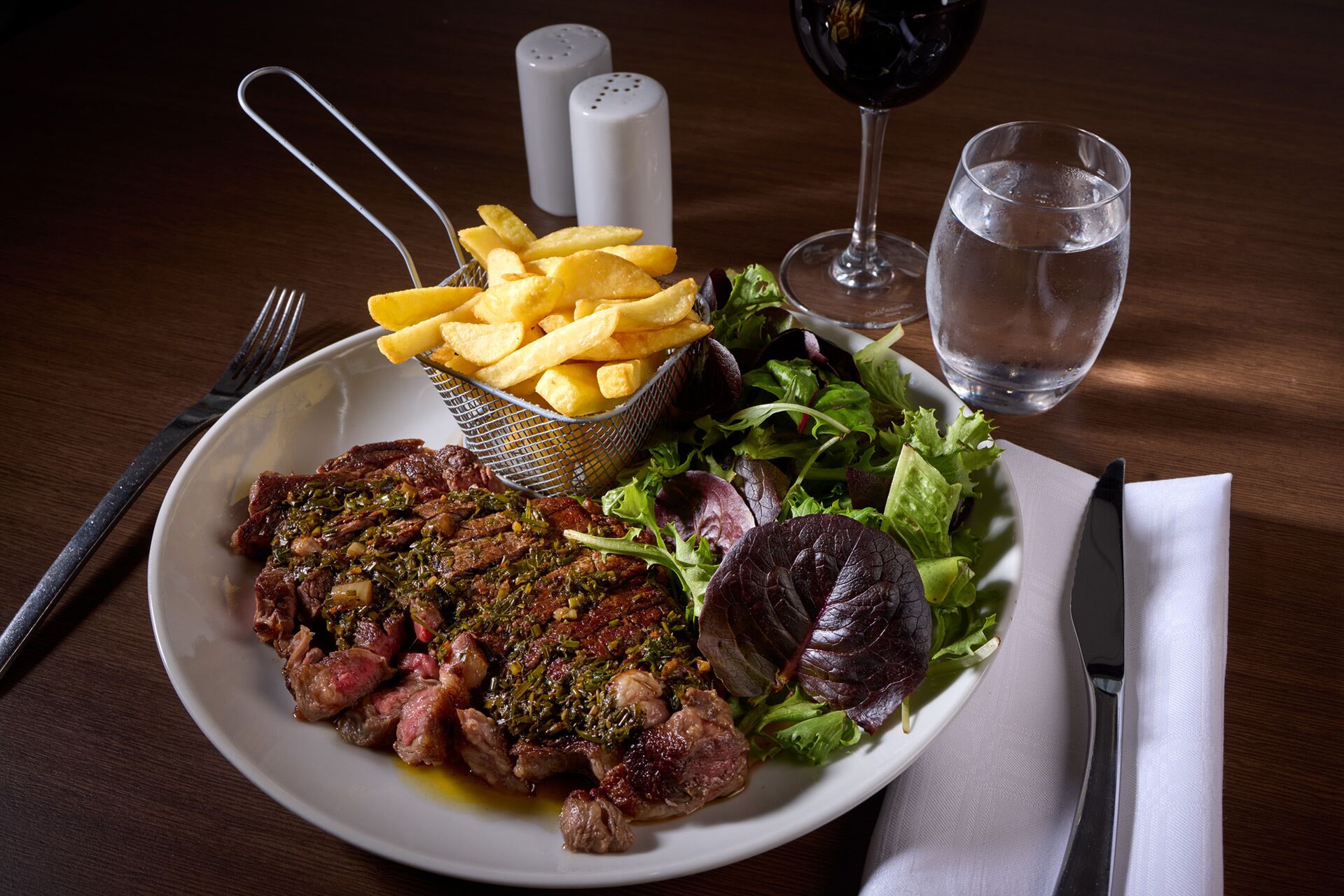 A plate with steak, fries, and salad, accompanied by a glass of red wine and a glass of water, on a wooden table.