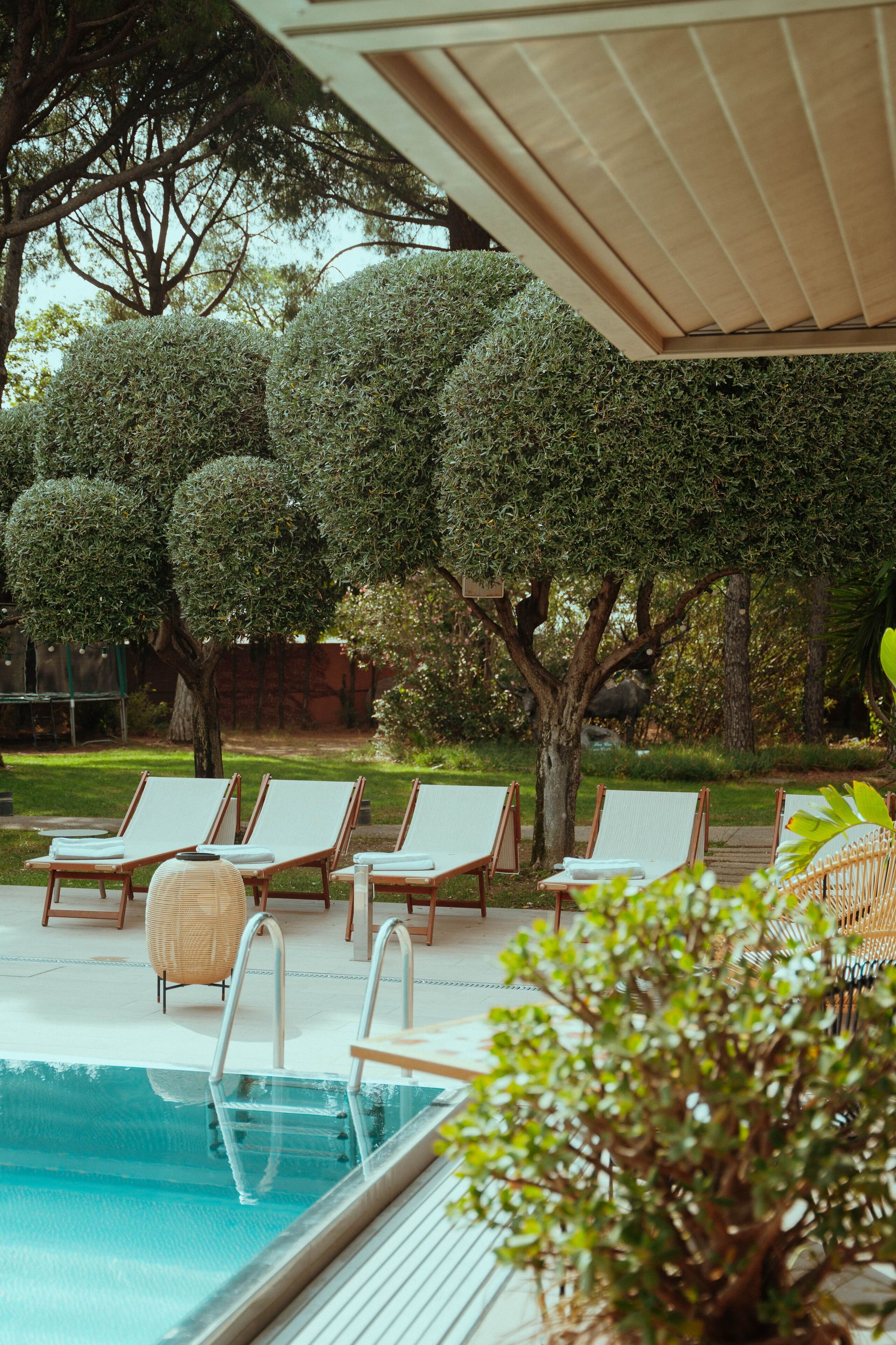 Uma área à beira da piscina com espreguiçadeiras, piscina e árvores bem aparadas.