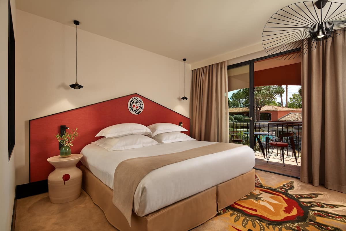 A hotel room with a large bed, a red headboard, a vase with flowers, a view of the outside balcony, and a ceiling fan.