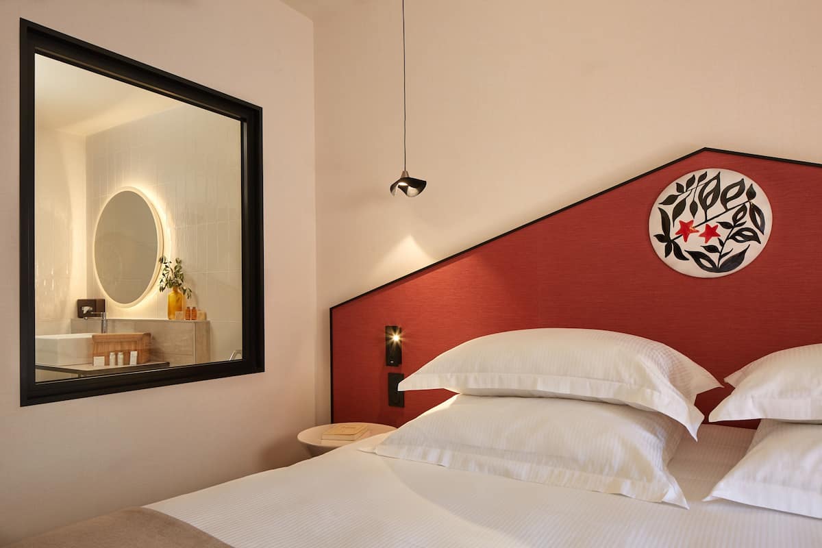 A hotel room with a large mirror, a bed with white linens, a red accent wall, and a wall-mounted light fixture.
