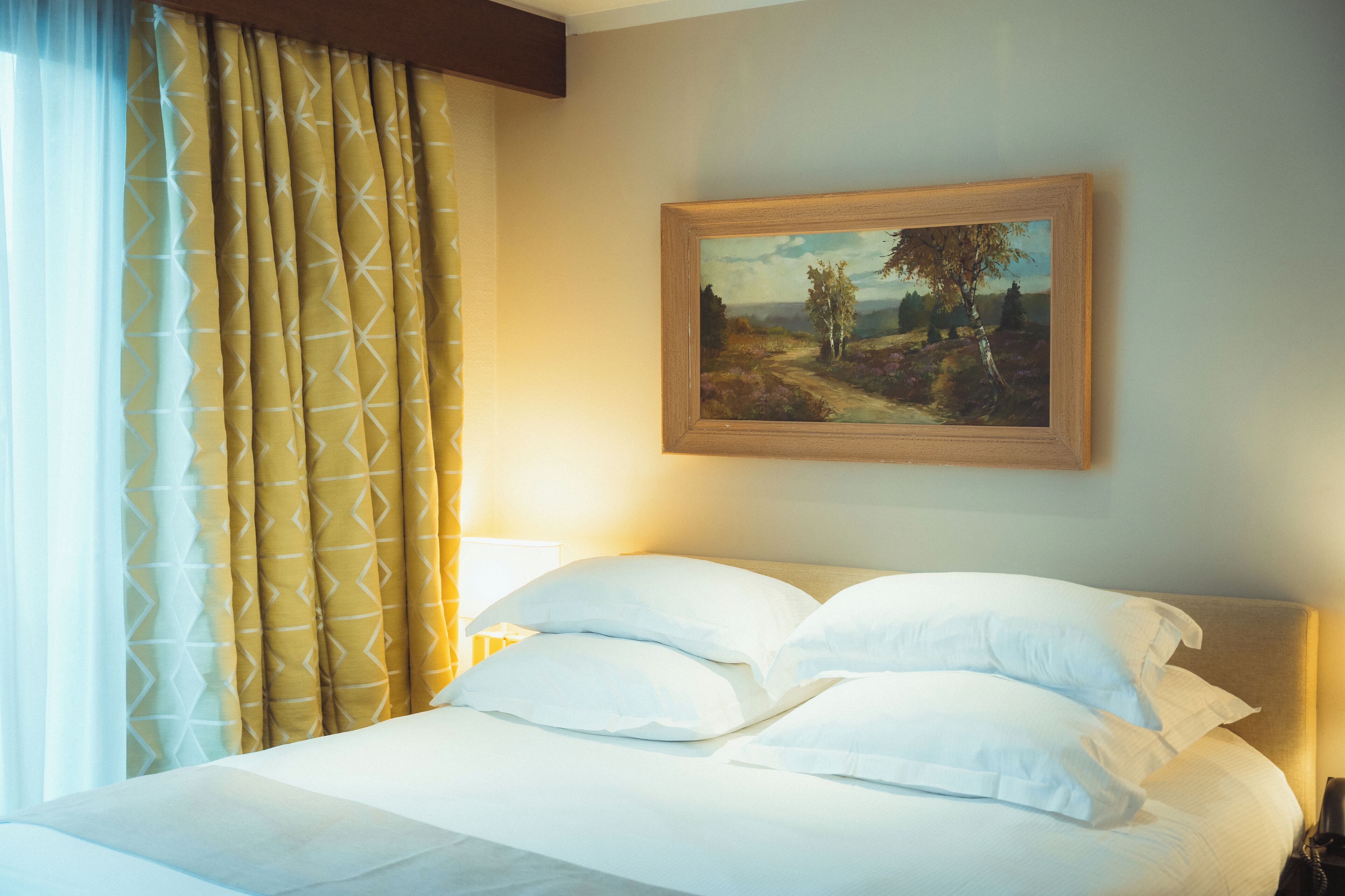Een hotelkamer met een bed, kussens, een schilderij aan de muur en gordijnen.