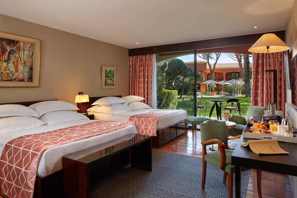 A hotel room with two beds, a dining area with breakfast, and a view of the garden.