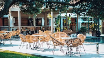 An outdoor seating area with wicker chairs and tables, surrounded by greenery and a pool.