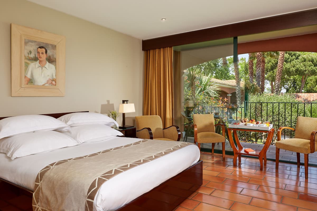 A hotel room with a large bed, a painting on the wall, a balcony with chairs and a table, and a view of greenery.