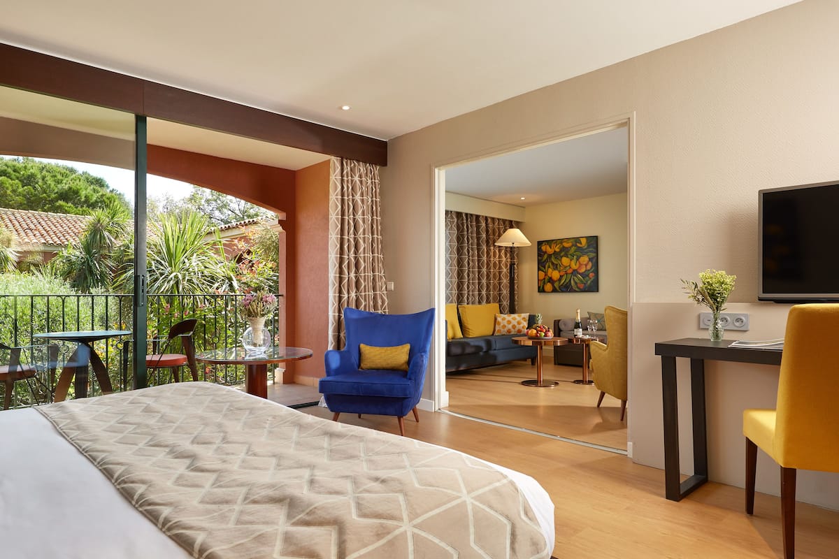 A hotel room with a bed, a blue armchair, a yellow chair, a desk with a TV, and a balcony with outdoor furniture.
