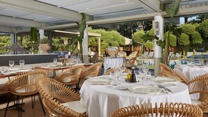 An outdoor dining area with wicker chairs and tables, surrounded by greenery and a pool.