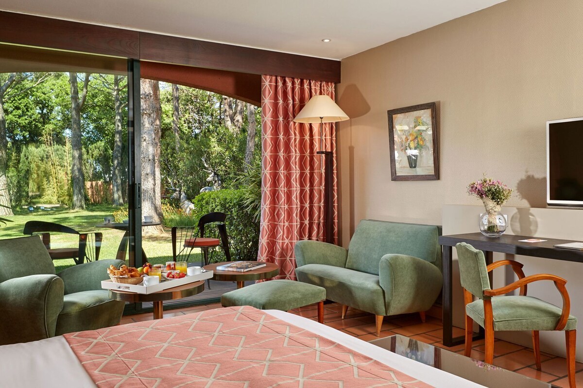 A room with a large window, a dining area with chairs and a table, a sofa, a TV, and a painting on the wall.
