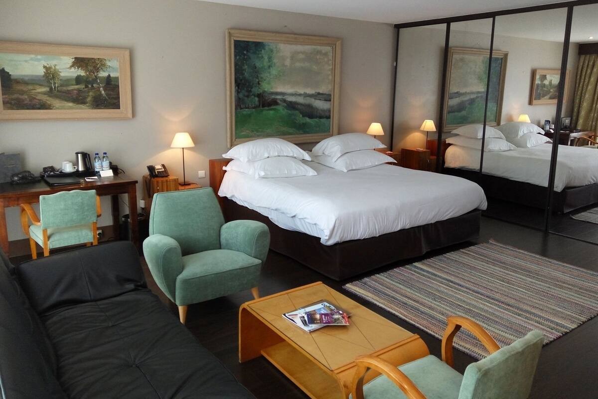 A hotel room with a bed, two armchairs, a sofa, a coffee table, a desk with a lamp, and a painting on the wall.
