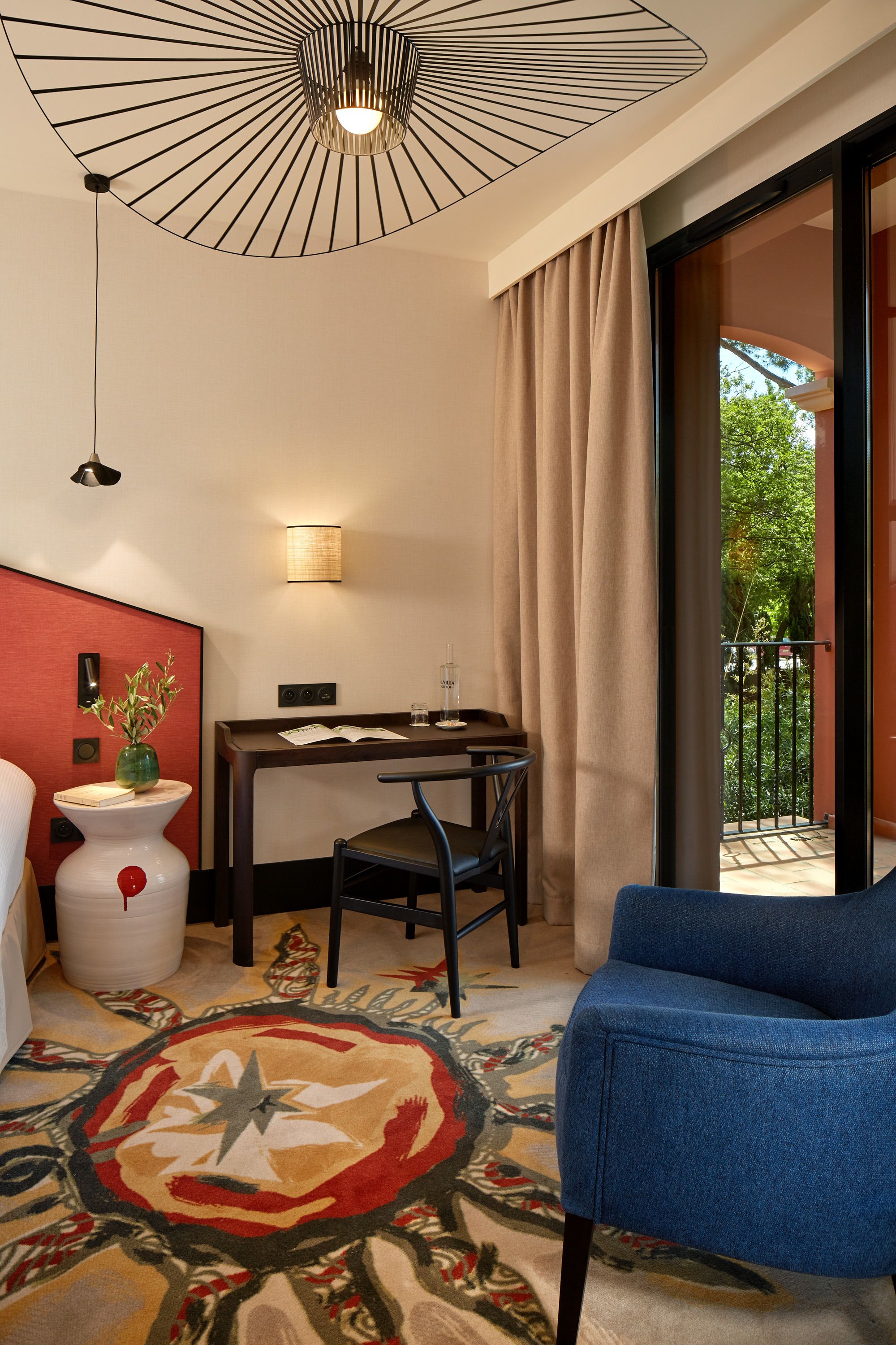 A room with a dining table, a blue chair, a decorative rug, a wall-mounted light, and a view of the outdoors.